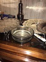 Silver plated servers, trays, bowl with glass insert, and silver plated shaker arranged on a wooden surface with vintage curtain in background