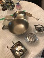 Overview of lot showing pewter bowls, lidded glass bowls, serving utensils, and square containers on padded surface under lighting.