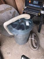 Metal garbage can with attached corrugated hose and coiled hose beside it
