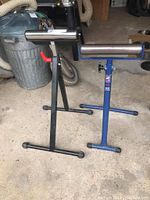 Two adjustable wood working stands with cylindrical rollers, one black and one blue, standing on a workshop floor.