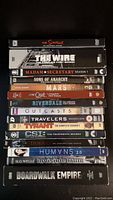 Stack of 15 DVD season cases showing titles including The Simpsons, The Wire, Madam Secretary, Sons of Anarchy, Mars, Riverdale, Outcasts, Travelers, Tyrant, CSI, Humans, The Invisible Man, Boardwalk Empire