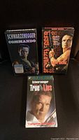 Front view of three sealed Arnold Schwarzenegger VHS tapes showing cover art for Commando, Raw Deal, and True Lies