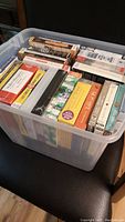 Photo of clear plastic bin filled with assorted sealed audiobook cases arranged vertically and horizontally showing colorful spines with titles.