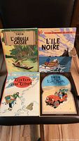 Four vintage French Tintin books laid out on a chair showing all front covers: L'Oreille Cassée, L'Île Noire, Tintin au Tibet, and L'Îlot Noir.