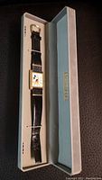 Full view of the wristwatch placed inside a light blue and beige presentation box with gold lettering on the inner lid.