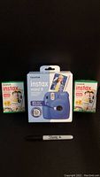 Blue Fujifilm Instax Mini 9 camera boxed, placed between two packs of Instax Mini instant film, with a Sharpie marker for scale.