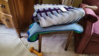 Two folded knitted wool afghans stacked on a small wooden stool, showing the purple and blue geometric patterned afghan on top and the solid light blue afghan underneath.