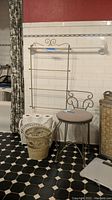 Photo showing metal towel holder rack mounted on wall, three wicker containers (wastebasket, magazine holder, hamper) plus metal vanity stool with beige cushion and bathroom scale on floor.
