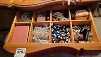 Open wooden drawer containing divided compartments filled with assorted costume jewelry and pins, plus a small pink quilted pouch.