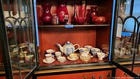 Full view of two open glass doors displaying cranberry glass items on top shelf and porcelain dishes on bottom shelf inside wooden cabinet.