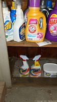 Various laundry products on wooden shelf, including All detergent, Arm & Hammer softener, Ajax cleanser, and spray bottles.