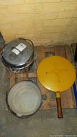 Overall view showing the Griswold cast iron pot with lid, white enamel pot with handles, and yellow Dansk frying pan with wooden handle lying on wooden pallets.