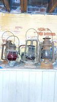 Five vintage hurricane lamps arranged on a wooden surface against a brick wall. Includes lamps with clear and red detail glass chimneys and metal frames showing rust and wear.