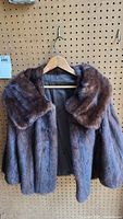 Front view of brown mink fur jacket hanging on a wooden hanger against pegboard background. The jacket has a large collar and visible soft fur texture.