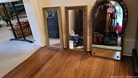 Three vintage framed mirrors on floor: two rectangular gold-tone framed mirrors and one tall arched mirror with floral etching.
