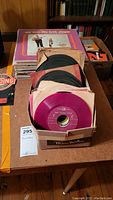 Photo showing multiple stacks of 45 RPM vinyl records in worn cardboard boxes and paper sleeves on a table.