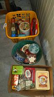 Overview of tins and trays including yellow plastic basket with spice tins, green basket with round tins, and cardboard box with additional tins and small bottles.