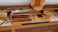 Photo showing vintage books stacked alongside ashtrays, yardsticks, pens, and a decorative plate with a sepia image of a building.