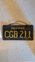 Front view of vintage 1967 California license plate with embossed yellow letters CGB 211 on black background showing hanging wire.