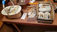 Photo showing two vintage wooden lunch baskets with decorated bakelite lids and three pairs of child's white shoes in boxes, placed on a wooden table.