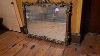 Full view of vintage rectangular mirror with ornate gold-toned frame on wooden floor leaning against wall.