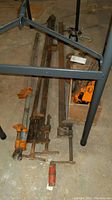 Photo showing several large metal bar clamps on floor with orange clamp heads, visible rust and wear