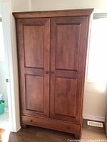 Front view of wooden armoire with two paneled doors and large drawer at the bottom on wood floor near window