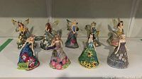 Front view of nine assorted Ashton Drake Gallery floral fairy figurines with wire loops for hanging, each with unique floral dress patterns and colors.
