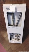 Boxed Taymor 4-piece bath hardware set, view showing full box upright with partial visibility of items inside including bar and mounting hardware.