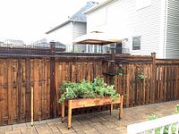 Fence mounted metal cat run, showing wire mesh enclosure attached on top of a wooden fence with a planter and patio visible below.