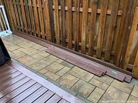 Three seven-foot composite deck boards laid flat against a wooden fence on outdoor stone pavers, brown color with textured wood grain surface.