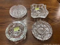 Top-down view of four distinct crystal ashtrays showing different cut patterns and shapes resting on wooden surface.