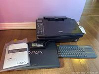 Sony VAIO laptop closed with manual and battery next to a black Epson Stylus NX510 printer and Logitech K400+ keyboard