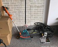 Photo showing the full lot: Worx weed trimmer, Yardworks leaf blower with power cord, garden rake, garden hose, boxes and small accessories on gray floor against brick wall.