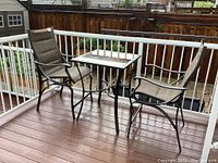 Patio table and two fabric cushioned metal frame chairs on a balcony deck, with wooden fence and neighboring houses in background.