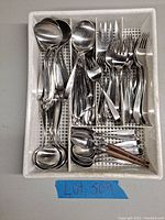 Top view of plastic tray holding assorted flatware: soup spoons, forks, knives, teaspoons, including some with wooden handles, showing quantity and condition with dust present.
