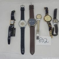 Full view of all 6 watches arranged horizontally on cloth background with lot number 802 card.