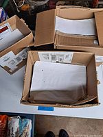 Three open cardboard boxes on a table, each filled with white wax-coated shipping sleeves. Boxes are worn and partially open so contents are visible.