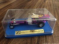 Side view of vintage purple metallic ZEE Cyclone dragster model inside clear display case on blue base with gold label.