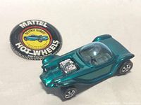 Photo of metallic teal vintage Hot Wheels Beatnik Bandit toy car with chrome engine on a white background alongside a Mattel Hot Wheels promotional button.