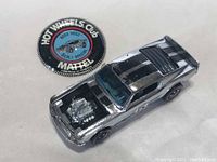 Die-cast car in silver finish with black stripes and redline wheels alongside round button badge