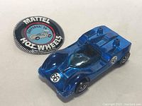 Top-down view of metallic blue Hot Wheels Chaparral 2G car positioned next to original round Mattel Hot Wheels Chaparral 2G button badge showing age wear.