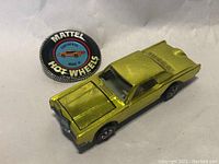 Yellow Mattel Hot Wheels Continental Mark III toy car with badge nearby on a white background. Shows front and side details of the car and the circular badge with graphics.