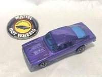 Front and side view of purple Hot Wheels Custom Barracuda car with redline tires and blue windshield along with the round Mattel Hot Wheels Custom Barracuda button with black background and yellow center.