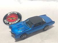Blue metallic Hot Wheels Custom Eldorado die-cast car with black roof, redline tires visible, and included black button with red car illustration and white lettering.