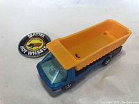 Photo of blue cab and yellow dump bed Hot Wheels dump truck and accompanying round Mattel Hot Wheels button/badge showing The Heavyweights Dump Truck branding