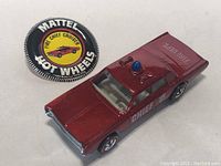 Photo showing Mattel Hot Wheels Fire Chief Cruiser die-cast car next to round button badge with Hot Wheels logo and Fire Chief Cruiser text.