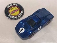 Top-down view of blue Hot Wheels Ford Mk IV car next to the round Mattel Hot Wheels Ford Mk IV badge