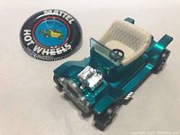 Close-up of the Hot Wheels Hot Heap toy car in teal with white interior, and the circular Mattel Hot Wheels Hot Heap button badge lying beside it.