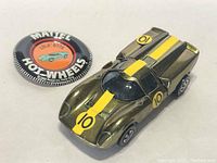 Fully visible image of gold Lola GT70 with yellow stripes, redline wheels, and original button showing Mattel Hot Wheels logo and Lola GT70 illustration.